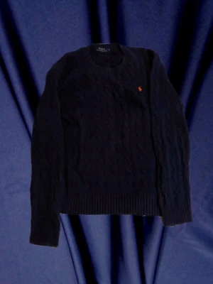 Mörkblå kabelstickad tröja Polo Ralph Lauren - Kabelstickad mörkblå tröja från Polo Ralph Lauren med rund halsringning och den klassiska broderade logotypen i orange på bröstet. Tröjan har långa ärmar och ribbade muddar vid ärmslut och nederkant. Perfekt för en stilren och tidlös look. I princip helt ny och har inte använts. Säljer för att dem var lite stor på mig. Skulle säga att den passar på personer från 180 till 190.