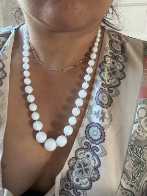 Vitt pärlhalsband med stora pärlor - Säljer ett snyggt halsband med vita, runda pärlor i olika storlekar. Pärlorna har en lätt räfflad yta som ger extra karaktär. Halsbandet har ett klassiskt och stilrent utseende och stängs med ett litet metallspänne. Perfekt för dig som gillar tidlös design.