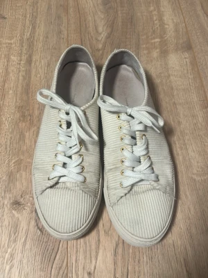 Vita sneakers i manchestertyg - Säljer ett par vita låga sneakers i manchestertyg med vita snören och guldfärgade öljetter. Skorna har en klassisk siluett och en vit sula som ger en clean look. Perfekta för dig som gillar stilrena och trendiga skor.