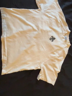 Vit t-shirt med stor tryck på ryggen - Vit t-shirt med svart grafiskt tryck på bröstet och en stor, detaljerad design på ryggen. Klassisk passform med korta ärmar och rund hals. Perfekt för dig som gillar streetwear och unika prints.