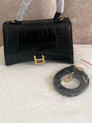 Svart läser glänsig handväska  - Snygg svart handväska med croc-mönstrat konstläder och guldfärgade detaljer. Väskan har ett stort B-spänne framtill och kommer med en avtagbar axelrem. Perfekt accessoar för att lyfta din outfit.