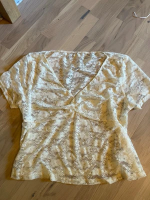 Beige spetsblus med v-ringning - Säljer en superfin ljus gul spetsblus med korta ärmar och v-ringad hals. Blusen är helt genombruten i spets och har en lätt rynkning framtill. Den är aldrig använd.