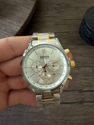 Silver och guld herrklocka från BOSS - Stilren herrklocka från BOSS med silverfärgad urtavla, gulddetaljer och länkarmband i rostfritt stål. Klockan har kronograf, datumvisning och tachymeter. Snygg kombination av silver och guld gör den till ett statement på handleden.