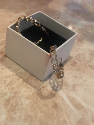 Guldiga hoops med strassdetaljer - Säljer ett par guldiga örhängen i hoop-modell med öppna länkar och gnistrande strass runt hela bågen. Örhängena har ett trendigt och lyxigt uttryck, perfekta för att lyfta vilken outfit som helst. Stift i metall och klassisk låsning. De har blivit lite rostiga men syns knappt när man har de på. Är öppen för prisförslag😊