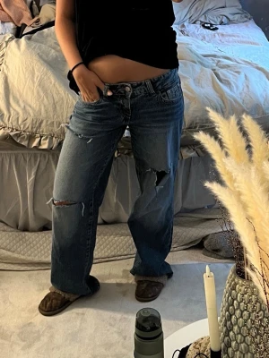 Blå slitna raka jeans - Säljer ett par blå raka jeans med slitningar och hål på knäna. Jeansen har en avslappnad passform och klassisk femficksdesign. Perfekta för en cool och avslappnad stil.
