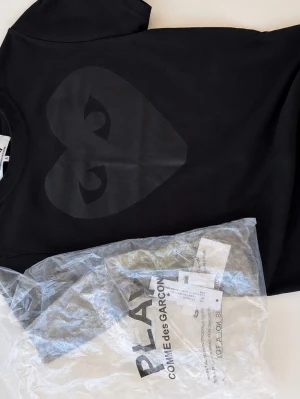 Svart Comme des Garçons PLAY t-shirt - Säljer en svart t-shirt från Comme des Garçons PLAY med det ikoniska hjärtmotivet i svart på bröstet. Klassisk rund hals och korta ärmar. Tillverkad i mjuk bomull för skön passform. Perfekt för dig som gillar stilrena plagg. Pris 399