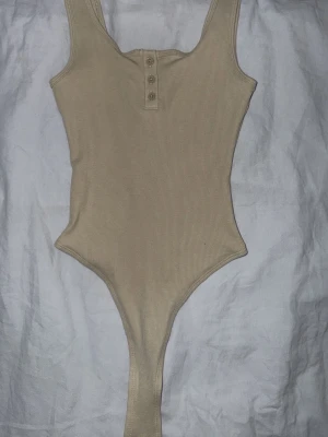 Beige ribbad body med knappar - Snygg beige body i ribbat material med breda axelband och tre dekorativa knappar framtill. Modellen har en klassisk skärning och är stretchig för en bekväm passform. Perfekt att styla till jeans eller kjol.