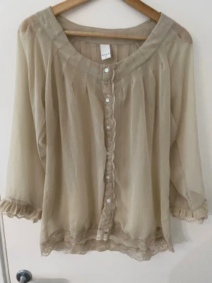 Blus - Blus från Vila i transparent, lätt material. Blusen har knappar framtill, spetsdetaljer längs knäppningen och nederkanten samt volang vid ärmslut.