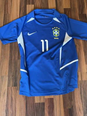 Brasilien borta tröja 2002 ( Ronaldinho ) - Snygg blå Brasilien fotbollströja från Nike med nummer 11 på bröstet. Tröjan har vita detaljer, rund hals och det klassiska brasilianska landslagsmärket . Perfekt för dig som älskar fotboll och vill sticka ut på planen. Även en av de bästa spelarna på ryggen, Ronaldinho.