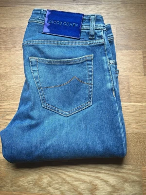 Jacob Cohën jeans - Säljer ett par klassiska blå jeans från Jacob Cohën med snygg kontrastsöm och en bakficka med unik sömdetalj. Jeansen har en smal passform och är tillverkade i mjukt denimtyg. Perfekta för dig som gillar stilrena och tidlösa plagg. Jeansen är inköpta på CareOfCarl och i väldigt bra skick, oerhört skön kvalité och passform. Modell Nick, storlek 31