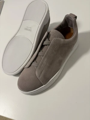 Grå sneakers från Zegna Triple Stitch - Orörd, först till kvarn, finns att testa i avenyn. Snygga grå sneakers från Zegna. Skorna har ovandel i mocka, elastiska band istället för klassiska snören och vit sula. 