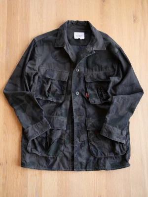 Camouflage Field Jacket - Säljer en grön och mörkgrå camouflage overshirt från Varsity med fyra stora fickor framtill och klassisk krage. Jackan är tillverkad i bomull och har knäppning med knappar. Perfekt för dig som gillar militärinspirerad stil och vill ha något som sticker ut.