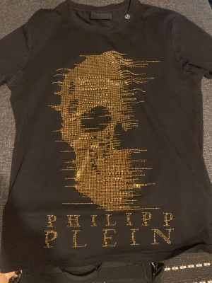 Svart Philipp Plein t-shirt med döskalle - Vet inte om äkta troligen inte. Svart t-shirt från Philipp Plein med guldiga nitar som bildar en stor döskalle framtill och logga både fram och bak. T-shirten har korta ärmar och en klassisk passform. Perfekt för dig som gillar edgy och lyxig streetstyle.