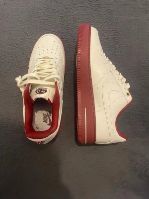 Nike Air Force 1 Low vit/röd - Nike Air Force 1 Low sneakers i vitt läder med röda detaljer och röd yttersula. Klassisk siluett med perforerad tåbox, vita skosnören och röd vadderad insida. Swoosh-logga på sidan och specialgrafik på plösen.