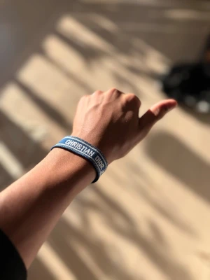 Blått Christian Dior tygarmband. Stort lager !!! - Snyggt blått tygarmband från Christian Dior med vit brodyr och texten 'CHRISTIAN DIOR' på framsidan. Armbandet har justerbar knytning med tofsar i ändarna och en cool, avslappnad look. Perfekt accessoar för att lyfta din outfit.