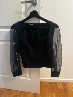 Svart topp med meshärmar från SHEIN - Trendig svart topp från SHEIN med transparenta meshärmar dekorerade med små pärlor. Toppen har en fyrkantig urringning och åtsittande passform. Perfekt för dig som vill ha något extra till outfiten.