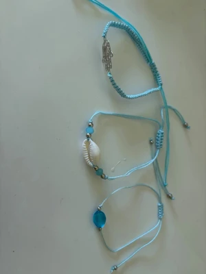 Set med turkosa armband - Säljer ett set med tre armband i ljusblått snöre. Ett armband har en silverfärgad handberlock, ett har en vit snäcka och små turkosa pärlor, och det tredje har en stor blå pärla i mitten. Perfekt för dig som gillar strandvibbar och boho-stil.