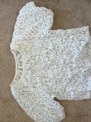 Vit spetsblus med kort ärm - Säljer en vit blus i spets med korta ärmar och rund halsringning. Blusen har ett genombrutet, blommigt mönster och är perfekt för dig som gillar en fin och luftig stil. Passar till både jeans och kjol.