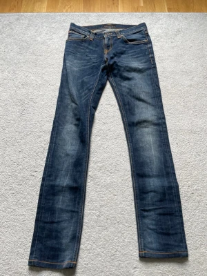 Blå raka jeans med slitningar - Säljer ett par klassiska blå jeans med raka ben och snygga slitningar. Jeansen har fem fickor, kontrastsömmar och en patch bak i midjan. Perfekta till en avslappnad stil och tillverkade i denim med skön känsla.