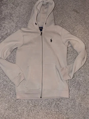 Beige hoodie från Polo Ralph Lauren - Snygg beige hoodie från Polo Ralph Lauren med dragkedja framtill och klassisk huva. Har två stora fickor på magen och den ikoniska Polo-loggan broderad på bröstet. Tillverkad i mjukt bomullsmaterial som känns riktigt skönt mot huden. Xs liten i storlek!