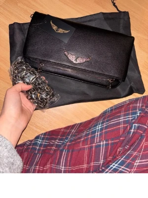 Svart handväska Zadig & Voltaire - Snygg svart handväska från Zadig & Voltaire i slätt konstläder. Väskan har ett coolt silverfärgat vingemblem på framsidan och dragkedja. Kedjeremmen ger en edgy vibe och väskan kommer med dustbag. Har taggen kvar och original påse.