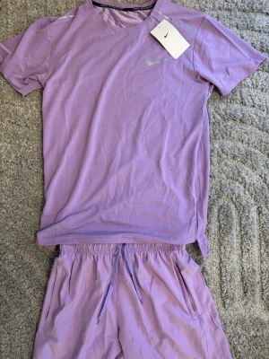 Lila träningsset från Nike - Säljer ett fräscht lila träningsset från Nike med matchande t-shirt och shorts. Setet är gjort i ett lätt och snabbtorkande material, perfekt för träning eller löpning. T-shirten har rund hals och Nike-logga på bröstet, shortsen har dragsko i midjan och fickor.