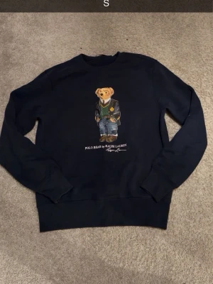Marin blå sweatshirt Polo Bear Ralph Lauren - Cool Marin blå sweatshirt från Ralph Lauren med den ikoniska Polo Bear-trycket på bröstet. Tröjan har rund halsringning, långa ärmar och ribbade muddar. Perfekt för dig som gillar streetwear och vill ha något klassiskt men ändå lekfullt. Ny pris 1500+, Helt ny tröja aldrig Andvänd säljer för 820