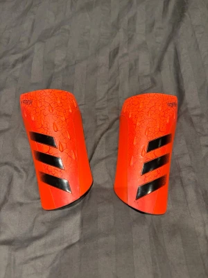 Röda benskydd från Adidas Predator - Säljer ett par röda benskydd från Adidas Predator-serien med svart mönster och de klassiska tre ränderna. Skydden har en sprucken texturdesign och är perfekta för fotbollsträning eller match. Lätta och smidiga att använda. Aldrig använda. OBS inget fodral