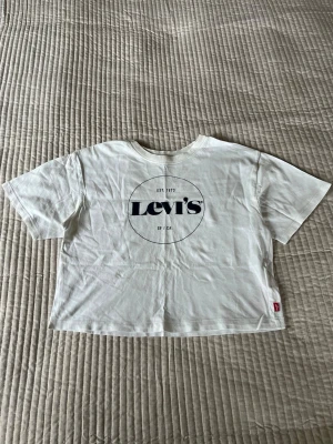 Vit croppad t-shirt från Levi's - Säljer en vit croppad t-shirt från Levi's med stor svart logga på framsidan. T-shirten har korta ärmar, rund halsringning och är tillverkad i mjuk bomull. Perfekt till jeans eller shorts för en avslappnad look. Snygg att matcha med ett par jeans eller en kjol. 