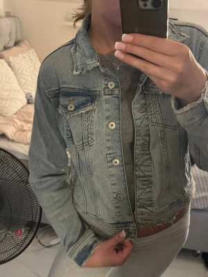 Ljusblå klassisk jeansjacka - Säljer en ljusblå jeansjacka med klassisk passform och silverfärgade knappar. Jackan har två bröstfickor med lock och knapp samt sömdetaljer framtill. Perfekt att slänga över en hoodie eller t-shirt för en chill look. Passar som S