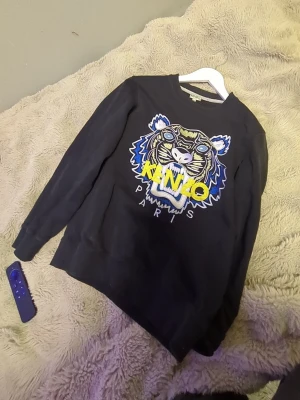 Svart Kenzo sweatshirt med tigertryck - Cool svart sweatshirt från Kenzo med stort broderat tigerhuvud och färgglada detaljer i blått, gult och vitt. Klassisk rund halsringning och ribbade muddar. Perfekt statement-plagg för dig som gillar streetwear och vill sticka ut.