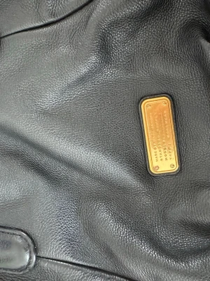 Svart handväska från Marc by Marc Jacobs - Snygg svart handväska i äkta läder från Marc by Marc Jacobs. Väskan har guldfärgade metalldetaljer och en klassisk rektangulär logoplatta framtill. Justerbar axelrem och innerfack med dragkedja för smart förvaring.