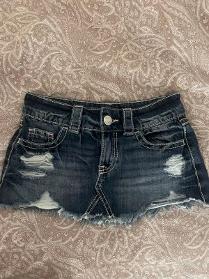 jeanskjol - Kort jeanskjol i mörkblå denim med slitna detaljer och fransig nederkant. Kjolen har klassiska fickor fram och bak samt vita kontrastsömmar. Perfekt för en avslappnad och trendig look.