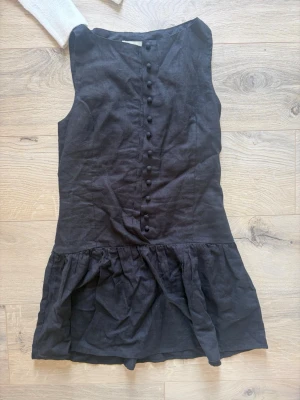 Svart linneklänning från H&M Xs - Säljer en svart ärmlös klänning från H&M i linneblandning. Klänningen har knappar framtill och en volang nedtill som ger en avslappnad look. Perfekt för varma dagar och enkel att styla med accessoarer.