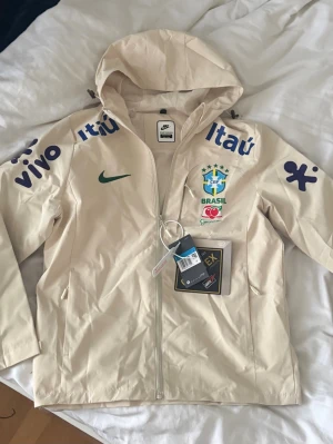 Nike Brasil vindjacka med huva - Beige Brasil windbreaker, helt ny och oanvänd, storlek M, hör gärna av vid frågor!