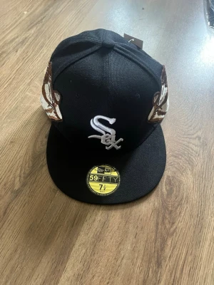 Svart New Era White Sox keps 59FIFTY - Säljer en svart New Era 59FIFTY keps med White Sox-logga framtill och broderade detaljer på sidorna. Baksidan har texten 'Jon Stan' i vitt broderi. Klassisk flat brim och cool streetwear-vibe. Materialet är bomull.