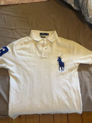 Vit pikéskjorta från Ralph Lauren - Vit pikéskjorta från Ralph Lauren med stor blå logga på bröstet och blå detalj på ärmen. Klassisk krage och knappar framtill. Perfekt för en clean och sportig look.