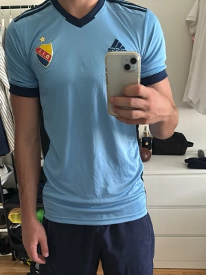 Ljusblå Djurgården matchtröja Adidas - Säljer en ljusblå Djurgården matchtröja från Adidas med mörkblå detaljer, klubbmärke på bröstet och nummer 2 på ryggen. Tröjan har korta ärmar, V-ringning och är tillverkad i ett lätt, ventilerande material perfekt för träning eller match.