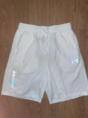 Vita shorts från Under Armour - Snygga vita shorts från Under Armour med elastisk midja och snörning. Shortsen har två sidofickor och logga i regnbågsfärg på ena benet. Tillverkade i ett lätt och svalt syntetmaterial, perfekta för träning eller varma dagar.