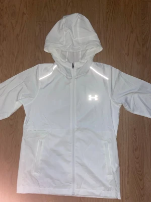 Vit vindjacka från Under Armour - Snygg vit vindjacka från Under Armour med huva och hel dragkedja framtill. Jackan har två sidofickor och reflekterande detaljer vid axlarna samt logga på bröstet. Perfekt för blåsiga dagar och sportiga outfits.