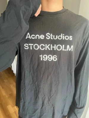Svart långärmad  Acne Studios äkta - Svart långärmad topp från Acne Studios med tryck 'Acne Studios STOCKHOLM 1996' på bröstet. Klassisk rund halsringning och relaxed fit. Perfekt till jeans eller kjol för en clean och stilren look.