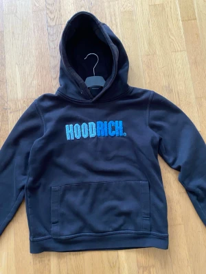 Svart hoodie från Hoodrich - Svart hoodie från Hoodrich med stor broderad logga i blå toner på bröstet. Klassisk känguruficka framtill och mjukt material som känns skönt mot huden. Perfekt för dig som gillar streetwear och vill ha en stilren men ändå iögonfallande look.