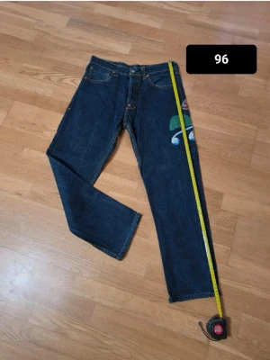 Evisu  - Unika mörkblå jeans med färgglatt broderi på ena benet. Klassisk femficksmodell med raka ben och kontrastsömmar. Jeansen har måtten 96 cm i längd och 38 cm i midja. Perfekta för dig som vill sticka ut med en personlig stil.