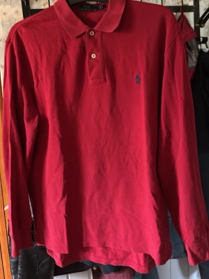 Röd långärmad piké från Ralph Lauren - Klassisk röd långärmad pikétröja från Ralph Lauren med krage och två knappar framtill. Tröjan har den ikoniska broderade loggan på bröstet och är gjord i mjuk bomull. Perfekt till jeans eller chinos för en clean look.