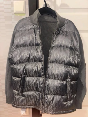 Grå dunväst från Dior - Snygg grå dunväst från Dior med ikoniskt monogrammönster över hela västen. Västen har stickade ärmar och krage i matchande grått, dragkedja framtill och två fickor. Perfekt för dig som vill ha en trendig och exklusiv look.