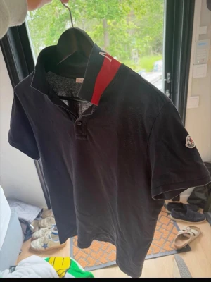 Moncler piké - Säljer en svart piké från Moncler med korta ärmar och klassisk krage. Kragen är röd med vit Moncler-logga på baksidan och pikén har en liten logotyp på ärmen. Tillverkad i bomull och har knappslå framtill. Pris kan diskuteras 