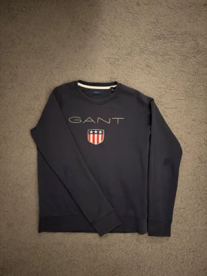 Mörkblå sweatshirt från GANT - Mörkblå sweatshirt från GANT med rund halsringning och tryckt logga samt ett sköldmärke med stjärnor och ränder på bröstet. Tröjan har långa ärmar och ribbade muddar vid ärmslut och nederkant. Perfekt för en avslappnad och stilren look.