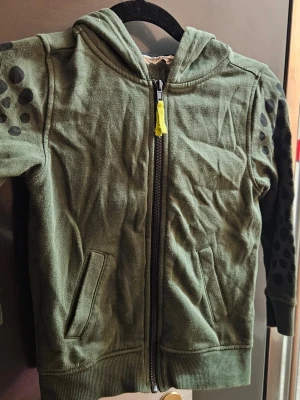 Grön hoodie med svarta prickar H&M - Grön hoodie från H&M med svarta prickar på ärmarna och dragkedja framtill. Två fickor på sidorna och huva. Tillverkad i mjukt bomullsmaterial, perfekt för en avslappnad och cool stil. Gul detalj på dragkedjan ger extra edge.