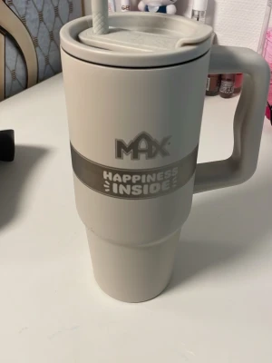 MAX Happiness Inside mugg med lock - Stor beige mugg från MAX med texten 'Happiness Inside' i grått. Muggen har ett praktiskt handtag, lock och sugrör, perfekt för drycker på språng. Tillverkad i plast och har en modern, stilren design.