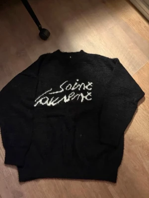 Svart Saint Laurent stickad tröja - Säljer en svart stickad tröja från Saint Laurent med vit logotyp i handskriven stil på bröstet. Tröjan har rund halsringning och långa ärmar. Materialet ser mjukt och fluffigt ut, perfekt för dig som gillar en trendig och avslappnad look.